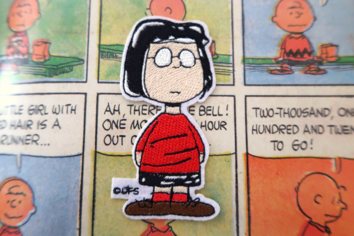 A die-cut embroidered patch of the character Marcie from Peanuts, wearing her iconic red sweater and black hair, placed on a vintage-style comic strip background.