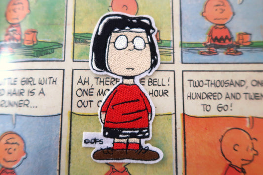 A die-cut embroidered patch of the character Marcie from Peanuts, wearing her iconic red sweater and black hair, placed on a vintage-style comic strip background.