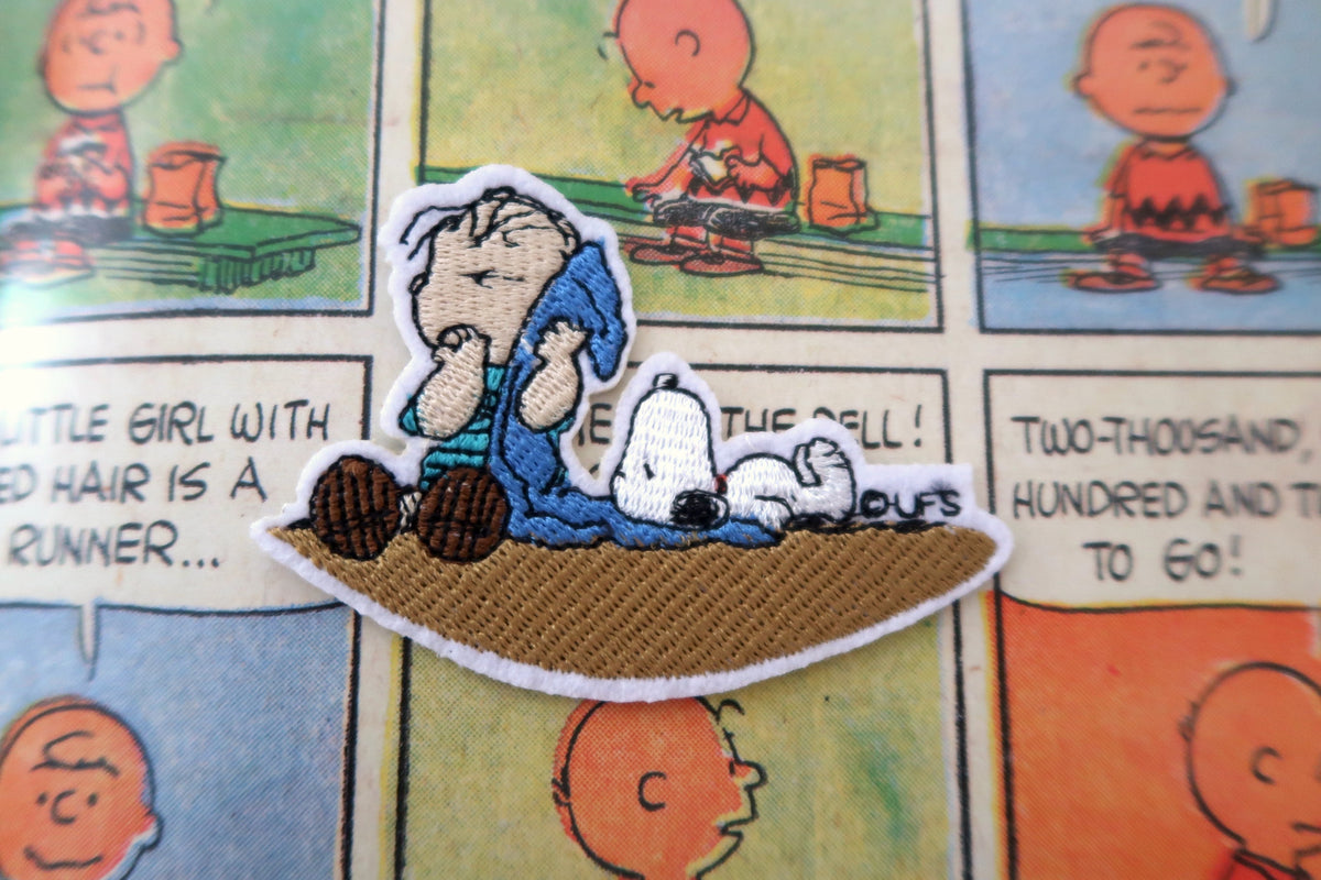 A close-up of a Peanuts embroidered patch featuring Charlie Brown and Snoopy lying on a comic book page. The patch shows CLinus van Pelt in blue pajamas with a blue blanket, Snoopy in white, and a brown base, with a white embroidered border.
