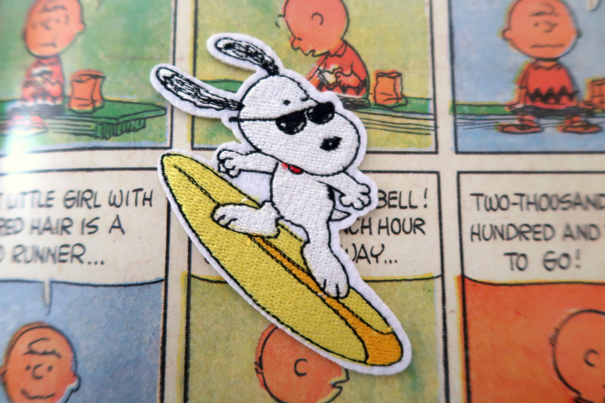 A close-up of a Peanuts Snoopy embroidered patch wearing sunglasses and riding a yellow surfboard, placed over a comic book page background.