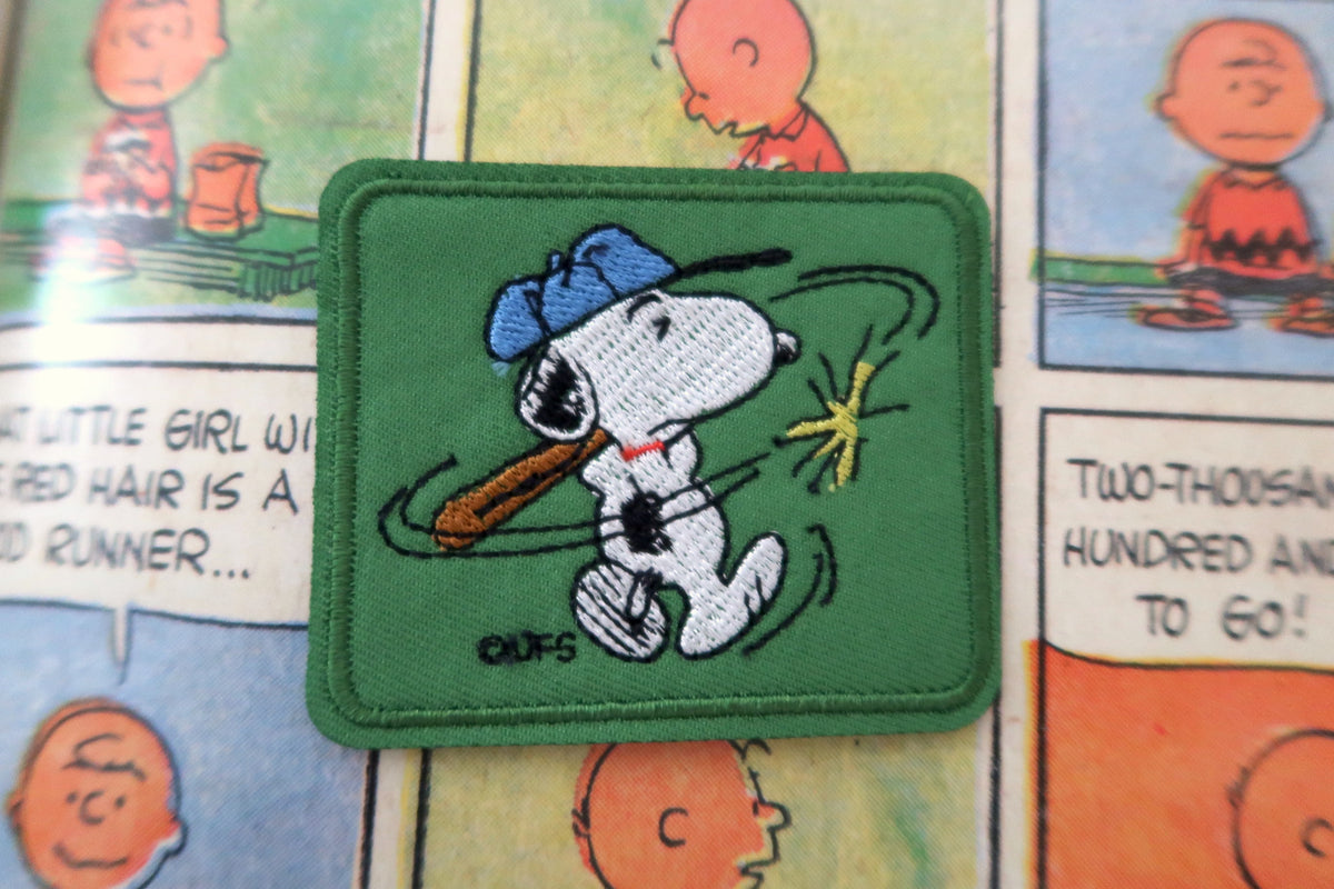 A square embroidered patch featuring Snoopy in a blue cap holding a brown bat, set against a green background, placed on top of a Peanuts comic book page.