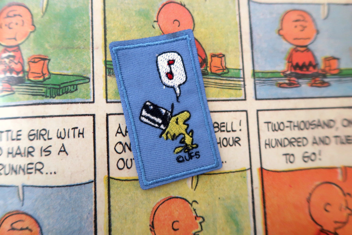 A blue embroidered patch featuring Snoopy and Woodstock with a speech bubble reading 'UFS' and a question mark, placed on a printed Peanuts comic page.