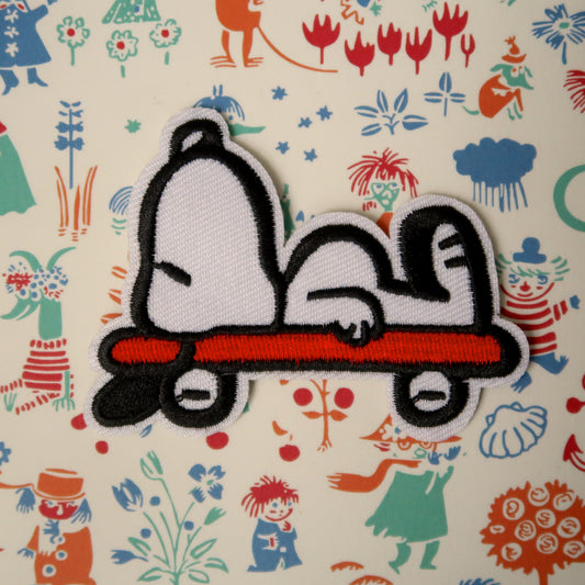A red and white embroidered patch featuring Snoopy from the Peanuts comics series sleeping on a skateboard.