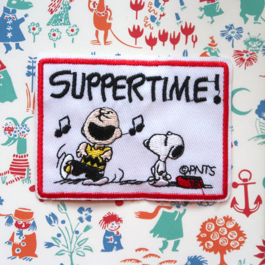 An embroidered patch featuring Snoopy and Charlie Brown from the Peanuts comic strip, with the text 'SUPPERTIME!' and musical notes.