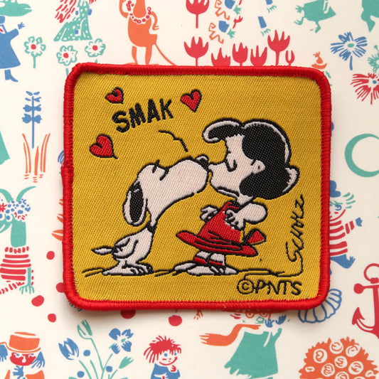 A red and yellow embroidered patch featuring Snoopy from the Peanuts comic strip kissing Lucy van Pelt.