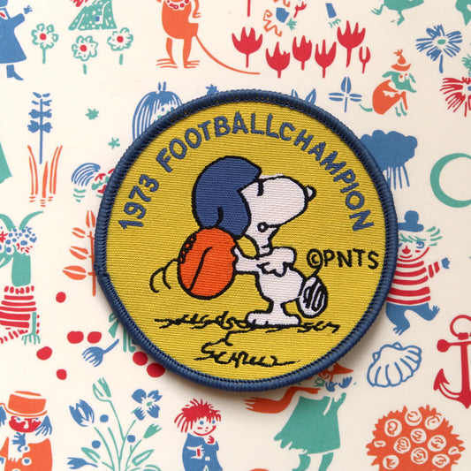 A yellow embroidery patch featuring Snoopy from the Peanuts comic strip with a football helmet and the text '1973 FOOTBALL CHAMPION'.