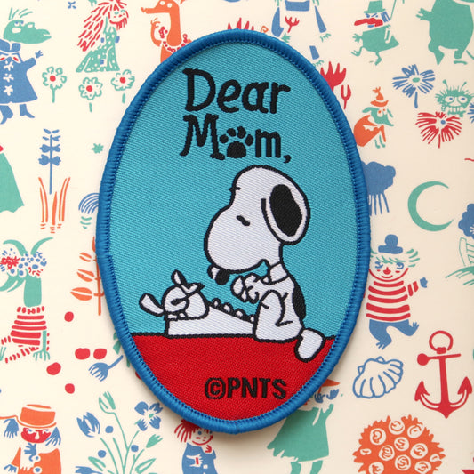 A blue embroidery patch featuring Snoopy from the Peanuts comic strip sitting at a white typewriter with the text 'Dear Mom' written in black.