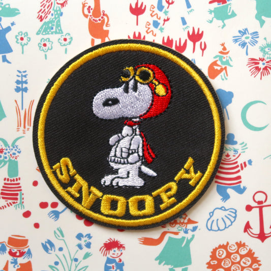 A round embroidered patch featuring the character Snoopy from the Peanuts comic strip, wearing a pilot hat and goggles, on a black background with yellow trim.