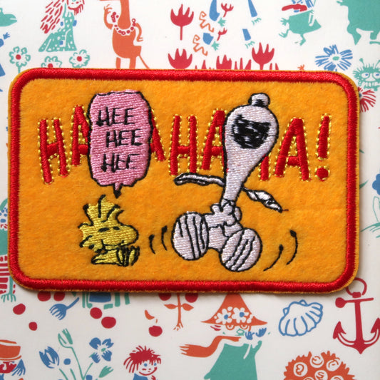 An embroidered patch featuring the character Snoopy from the Peanuts comic strip, with the text 'Ha Ha Ha Ha!' and an illustration of Snoopy and Woodstock.