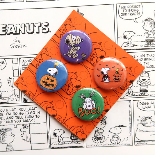 A set of four vintage style Halloween themed pin badges featuring characters from the Peanuts comics, placed on an orange background with a comic strip pattern.