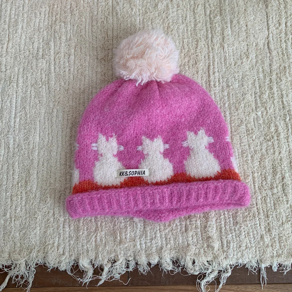 Pink knit hat with white cat design and pom-pom on a textured surface