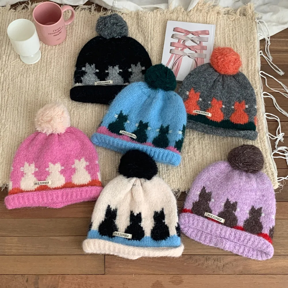Collection of colorful knit hats with fair isle cat designs and pom-poms on a textured surface