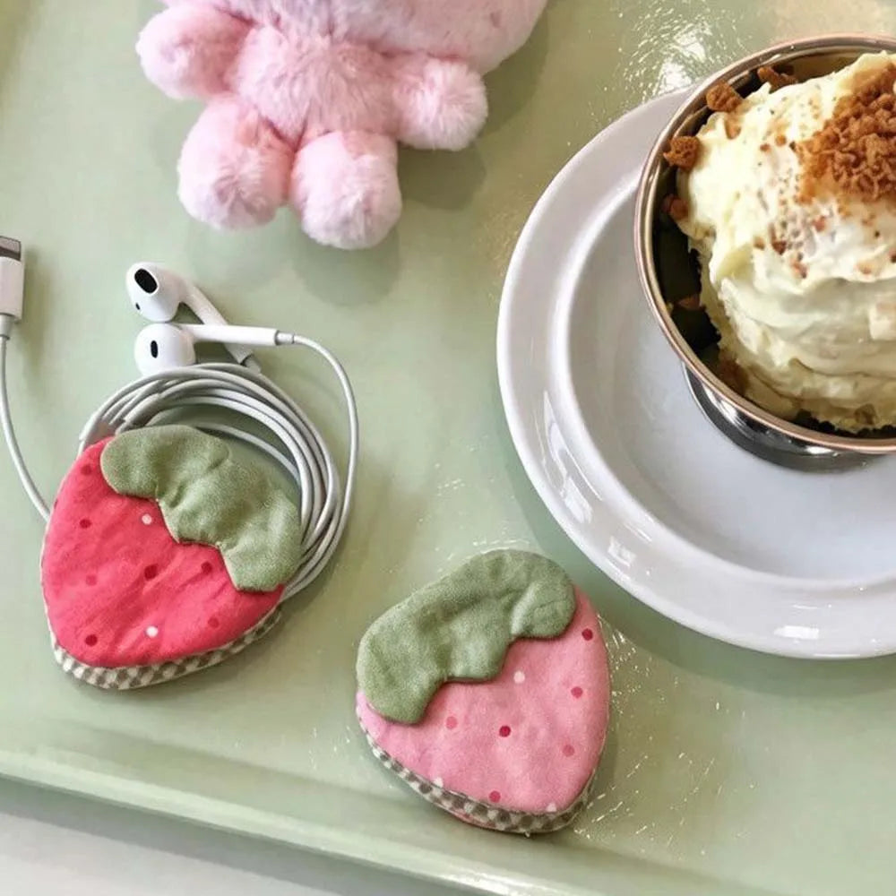 Two strawberry-shaped phone cable organizers in pink and green fabric, shown holding white wired earphones on a pale green tray. A dessert in a metal cup sits on a white plate nearby, with a pink plush toy partially visible at the top.