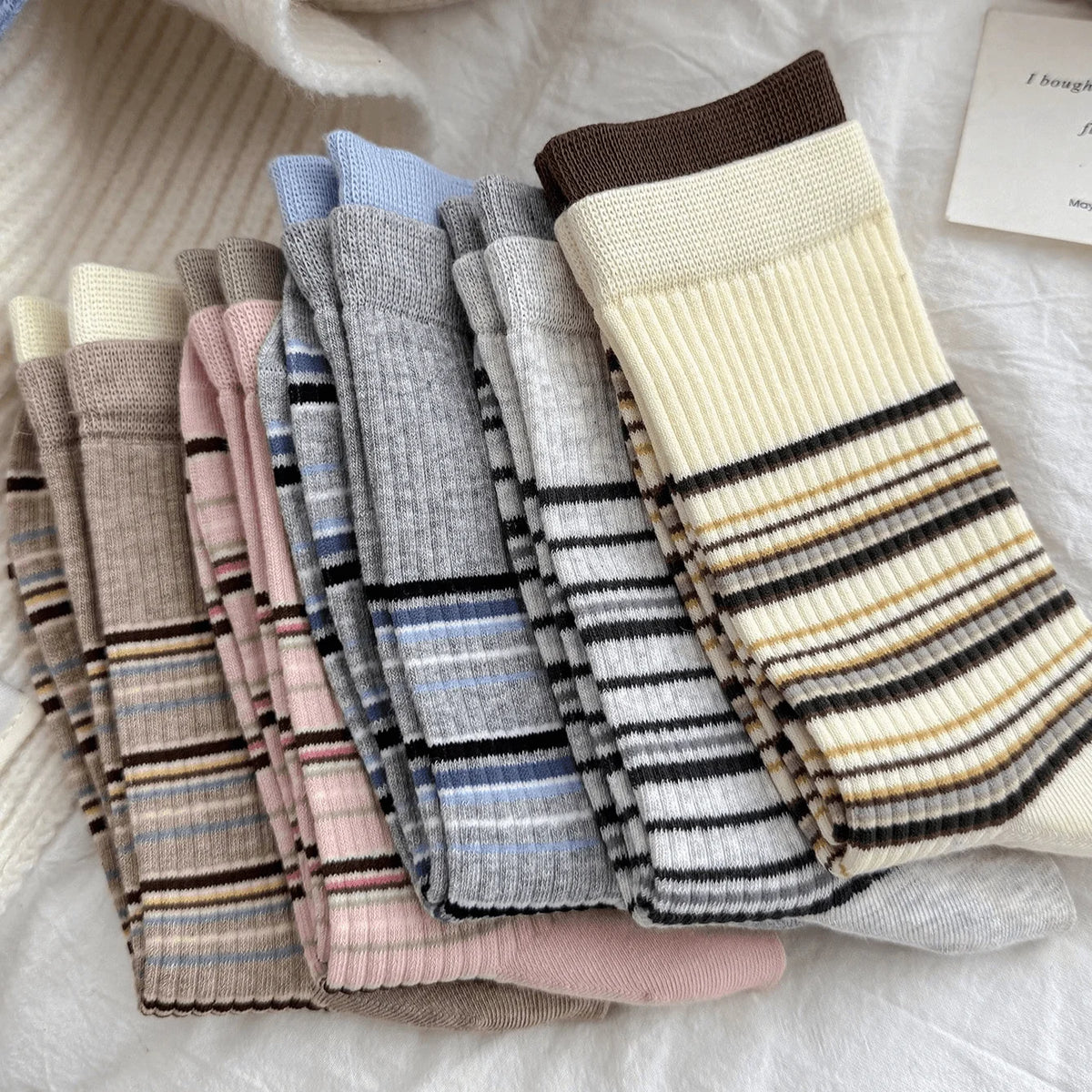 A set of six ribbed knit crew socks in various colors and stripe patterns, arranged in a fan shape on a soft white textured fabric background.