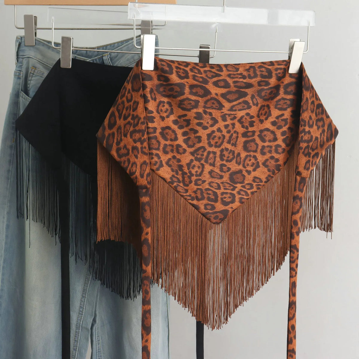 A leopard-print fringed wrap top hanging on a clear display hanger, shown alongside other garments on metal hangers against a plain light background.