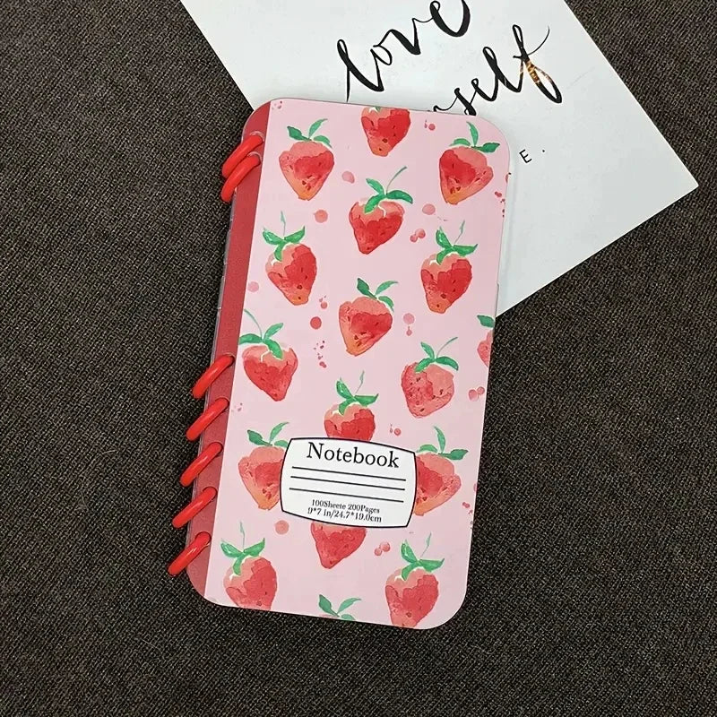 A pink notebook shaped iPhone Case with a strawberry pattern and red elastic loops, placed on a dark gray textured surface with a white card partially visible behind it.