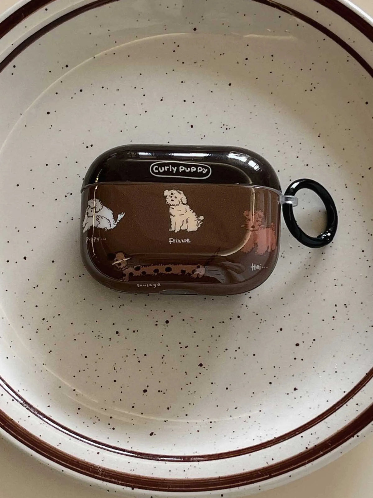 A brown AirPods case cover with dog illustrations and a black ring clip, placed on a speckled off-white ceramic plate.