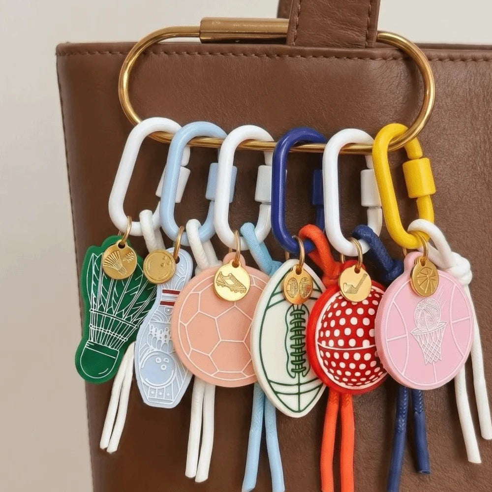 A close-up of a brown leather bag with a gold-tone ring holding six colorful sports-themed keychains. The keychains include designs for badminton, billiards, soccer, football, basketball, and ice cream, each with a round themed tag and dangling cords.
