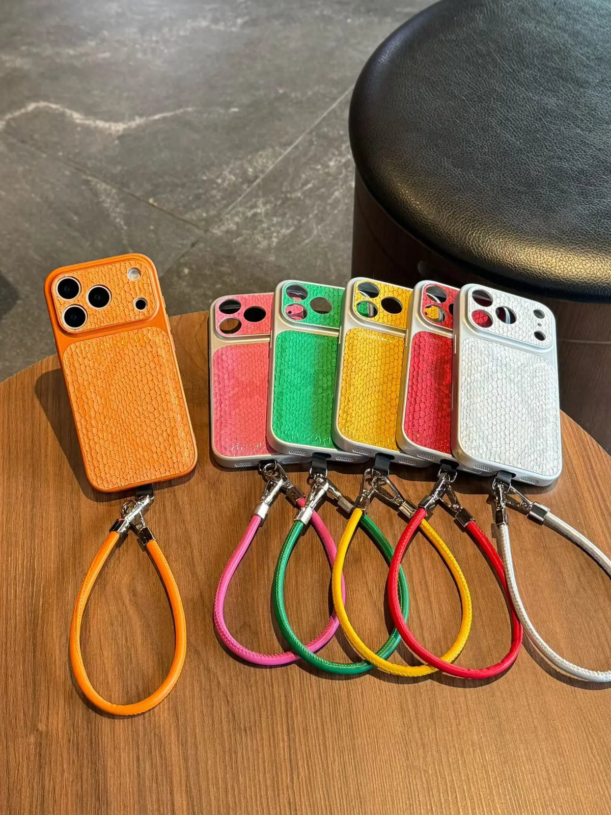 Six textured leather phone cases in orange, pink, green, yellow, red, and white, each with a matching wrist strap, arranged on a wooden tabletop.