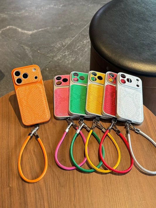 Six textured leather phone cases in orange, pink, green, yellow, red, and white, each with a matching wrist strap, arranged on a wooden tabletop.