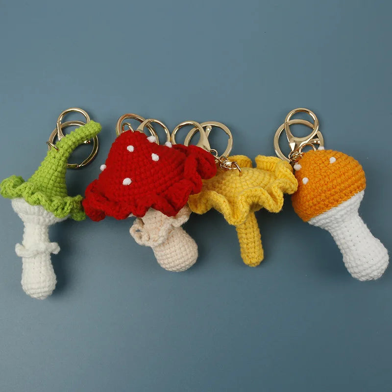 Four crocheted mushroom keychains in green, red, yellow, and orange, arranged in a row on a solid blue-gray background.