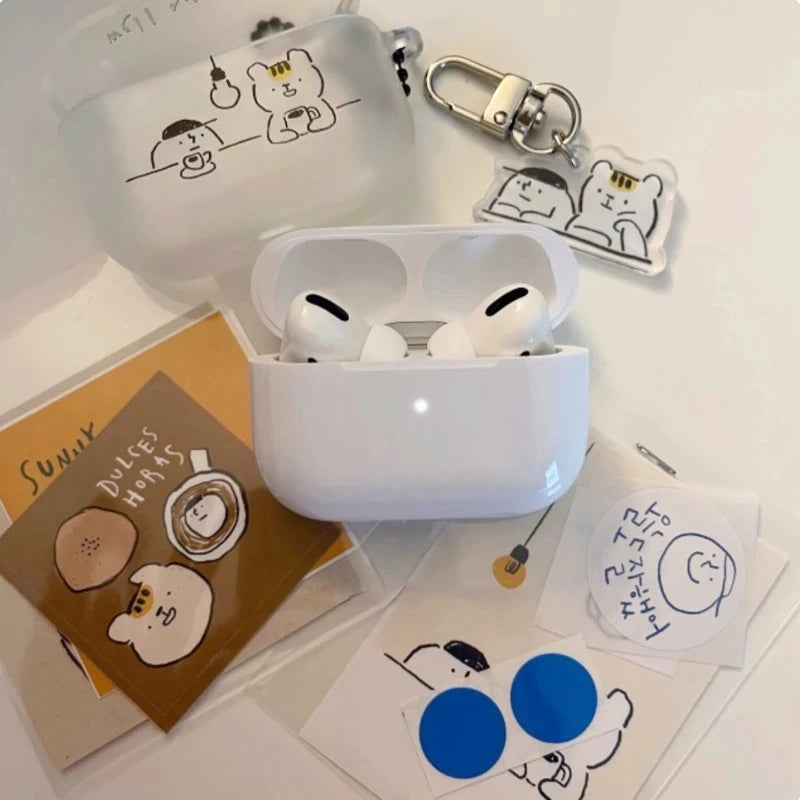 A white AirPods case with a translucent silicone cover featuring a cute cartoon bear drawing, shown open with AirPods inside. The set includes matching accessories: a keychain, a small pouch, and several stickers/cards with the same cartoon character on a white background.