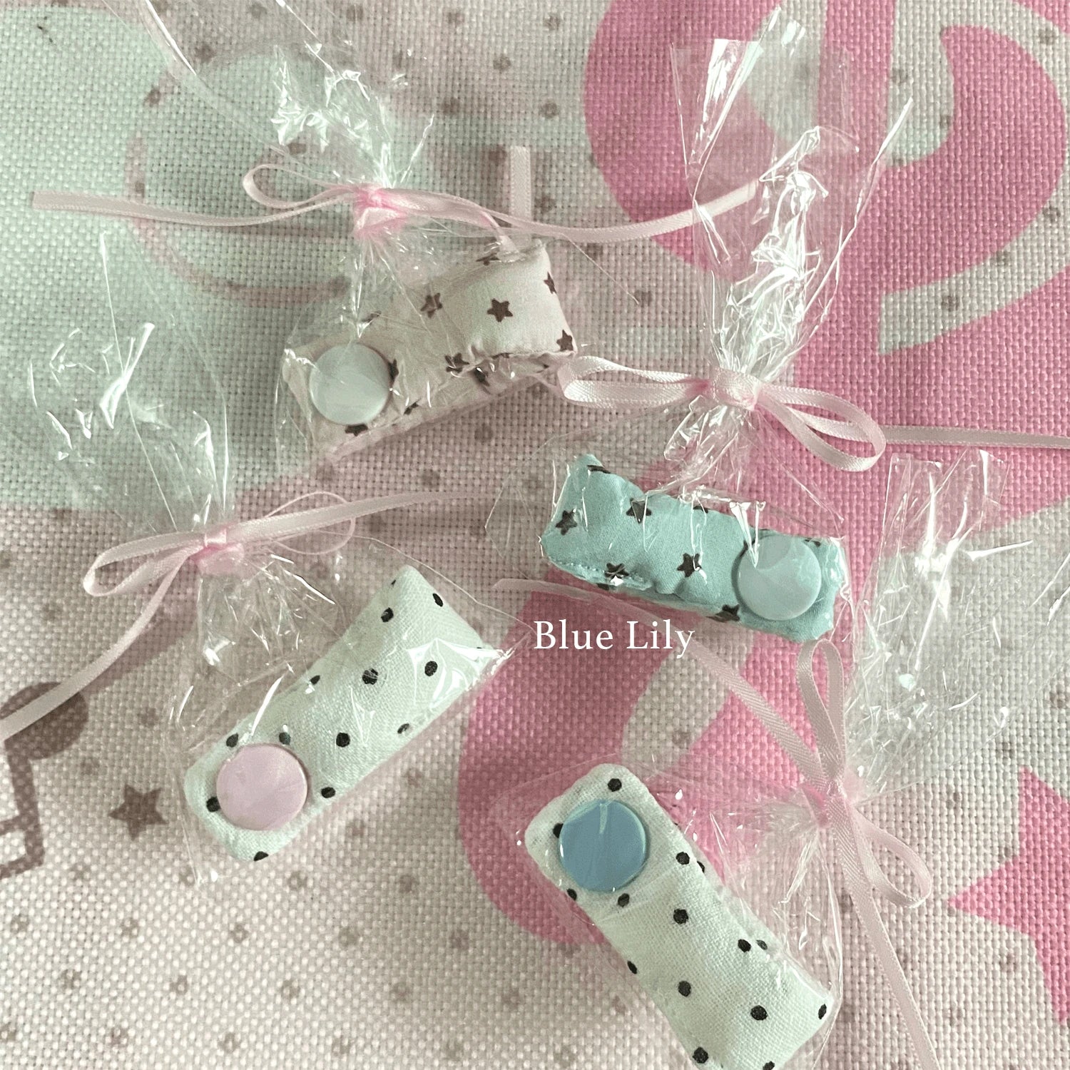 Four small fabric-covered snap buttons or charm pieces wrapped in clear cellophane bags with pale pink ribbons, arranged on a pink-and-white patterned fabric background. The text 'Blue Lily' appears over the center.