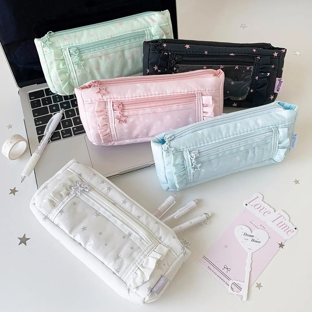 Four fabric pencil cases in pastel shades of mint green, black with pink stars, light pink, and light blue, arranged on a white desk next to a laptop, pens, and a card.