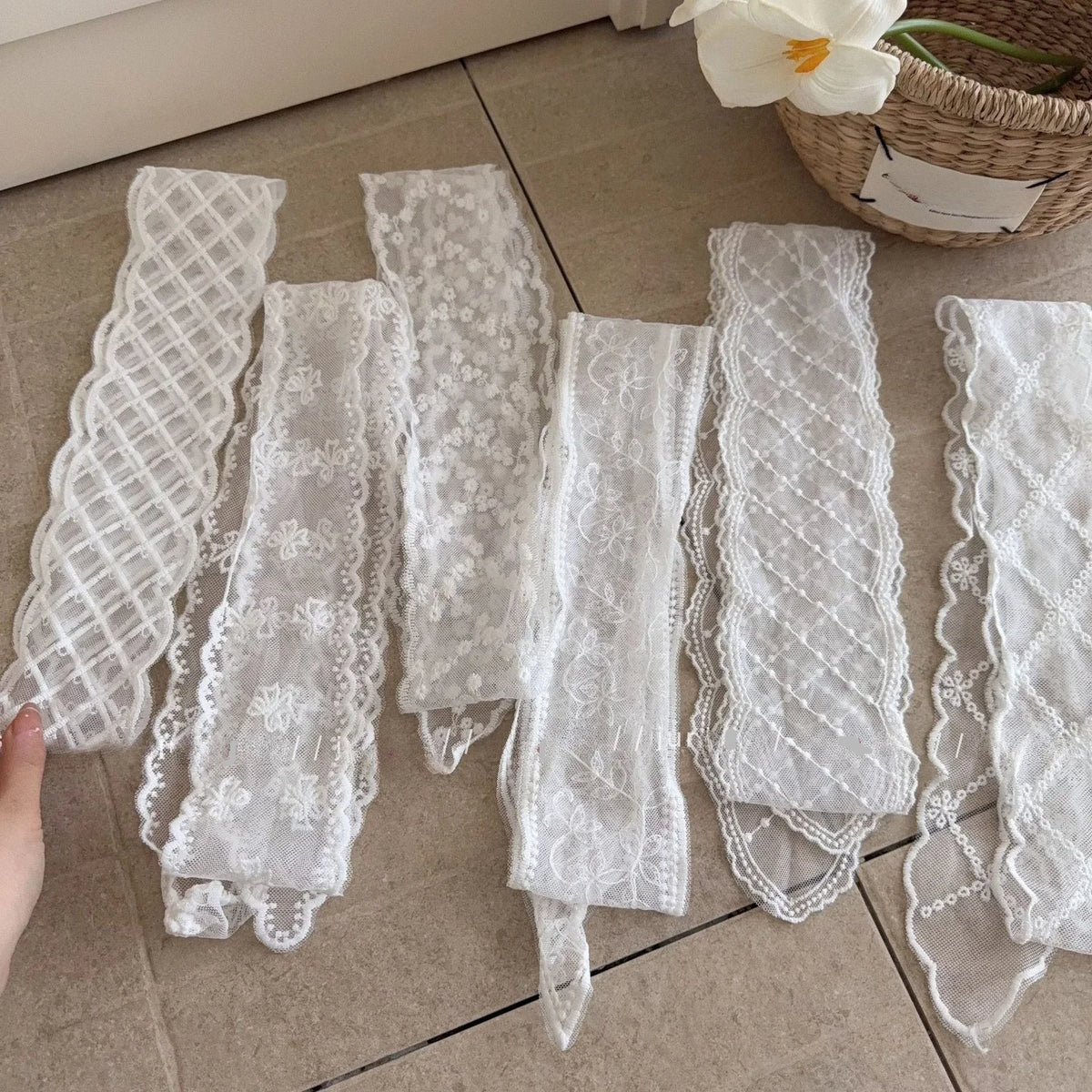 Six white lace trim strips of varying patterns and scalloped edges are laid out on a beige tiled floor, with a hand holding one strip on the left and a woven basket with a white flower in the upper right corner.