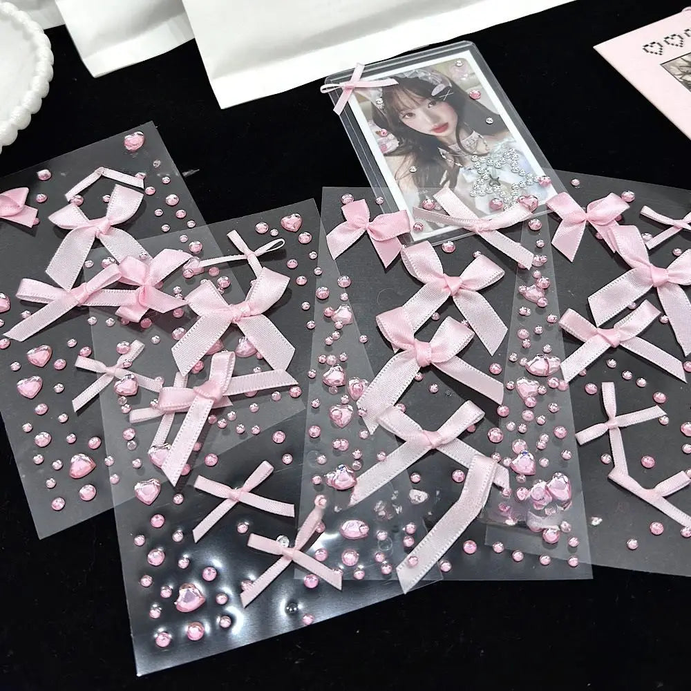 Several clear decorative cards or envelopes decorated with pink satin bows, pink rhinestones, and small pink heart embellishments, arranged on a black surface. One card near the top right shows a printed anime-style portrait.