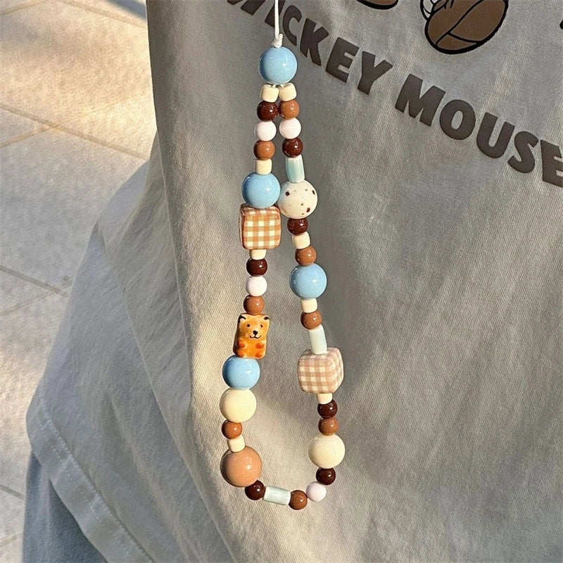 A beaded phone strap with pastel blue, brown, cream, and checkered accents hangs against a light gray fabric backdrop with a printed graphic.