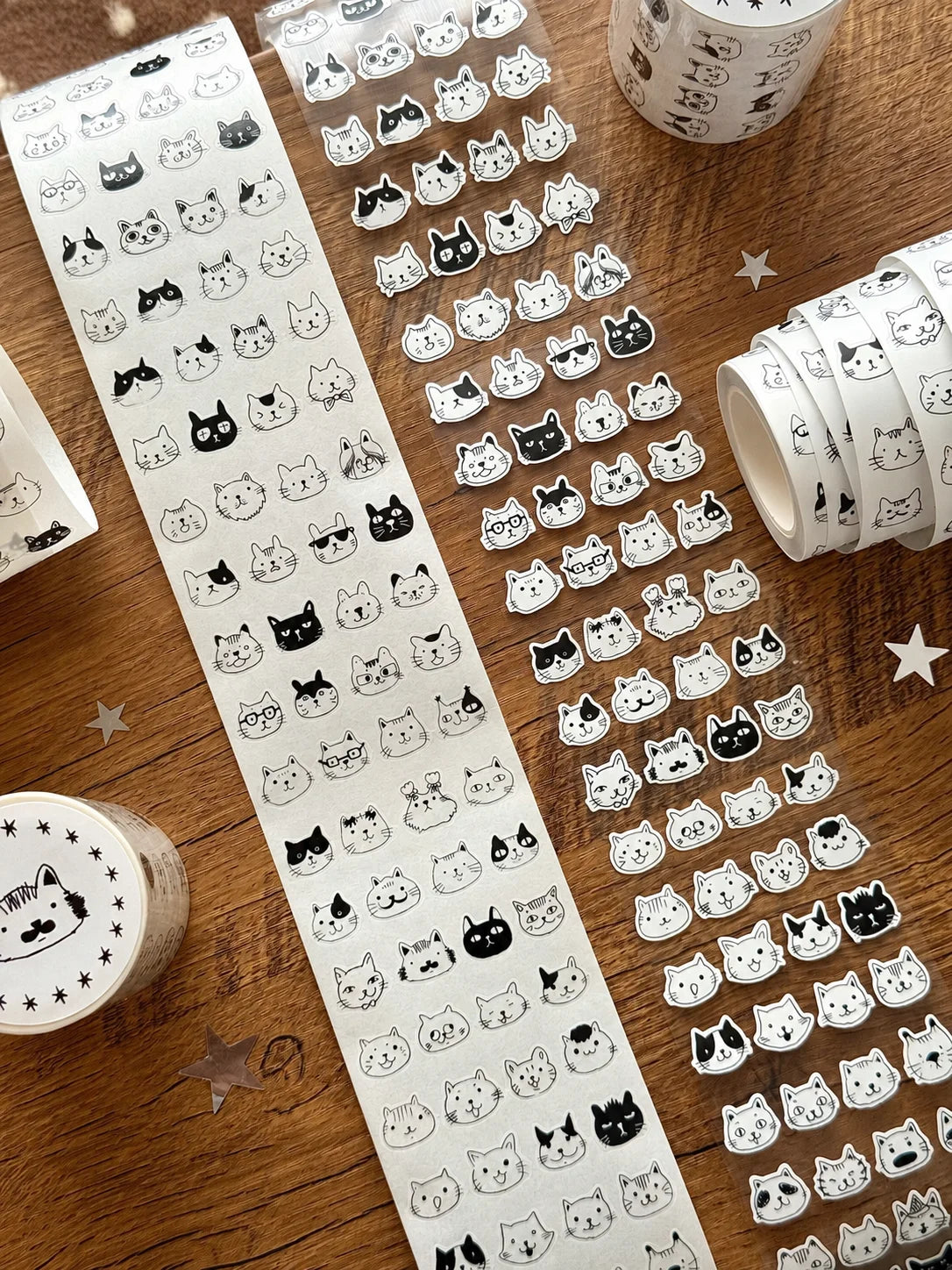 Washi tape rolls and unrolled strips featuring cute black and white cat face illustrations, arranged on a wooden tabletop with star confetti.