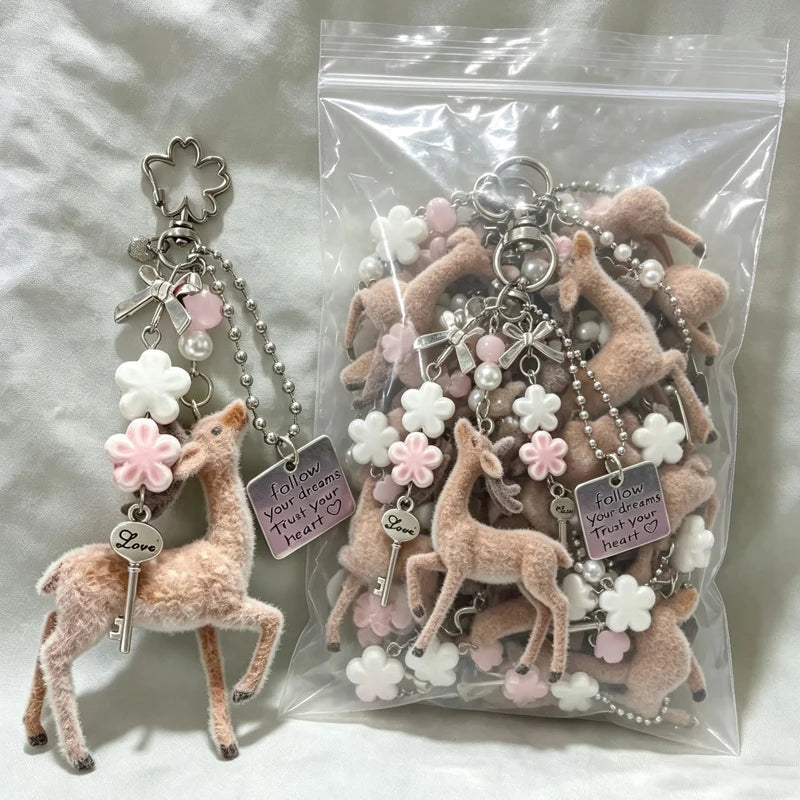 A plush deer keychain with silver charms, beads, and a key tag is displayed next to a clear plastic bag filled with multiple matching keychains on a white fabric background.
