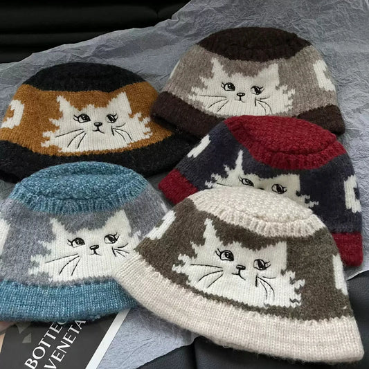 Five knitted cat-face beanie hats arranged on a crumpled gray fabric surface. The hats have different color combinations including black and brown, mustard and brown, light blue and gray, red and navy, and cream and brown.