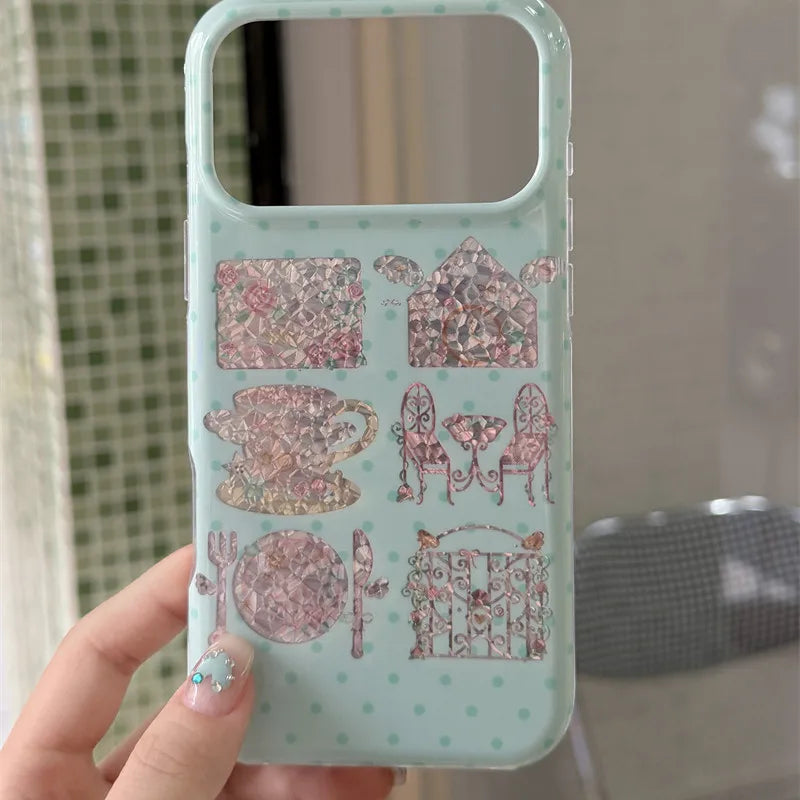 A hand holds a light mint green phone case decorated with pink and rose gold glittery illustrations of a teacup, teapot, chairs, and a gate, set against a blurred indoor background.