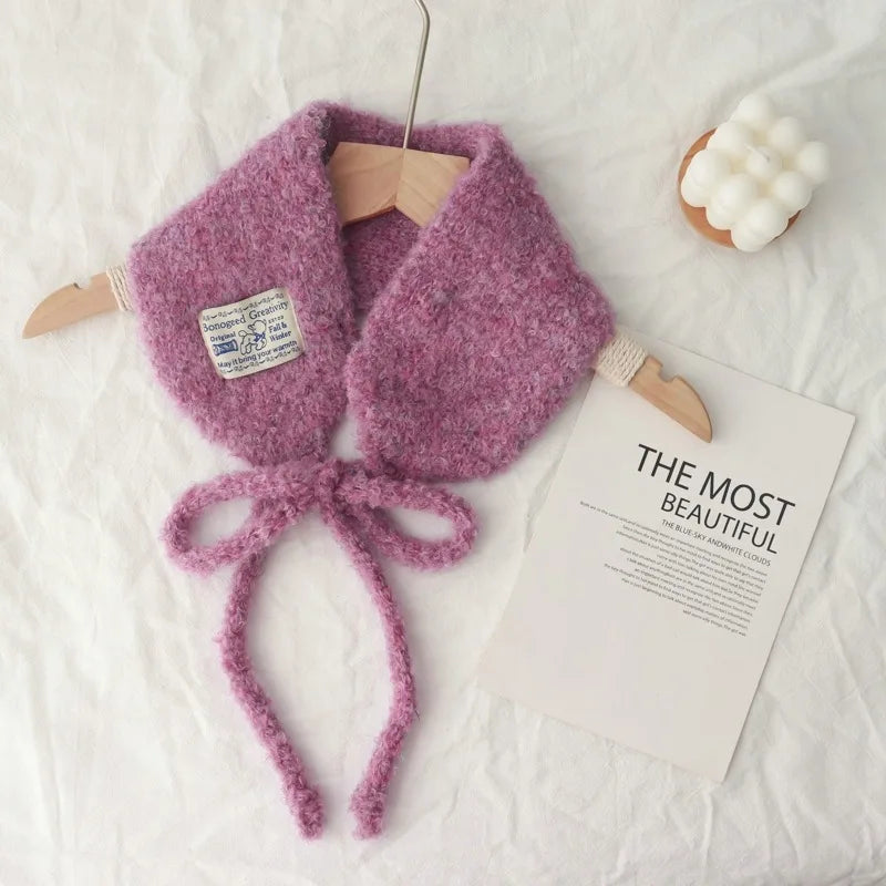 A fuzzy knit wrap or scarf in dusty pink, styled on a white fabric background with a wooden hanger, a decorative card, and a small white flower prop.