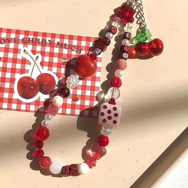 A cherry-themed beaded necklace with red and white beads, a pink polka-dot die charm, and a green cherry accent, displayed on a beige surface next to a red-and-white gingham card featuring cherries.