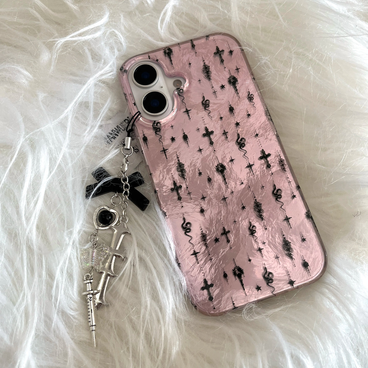 A pink phone case with a black gothic cross and skull pattern, featuring a dangling charm with a black ribbon bow, silver crossbones, and a small skull accent, displayed on a white faux fur background.