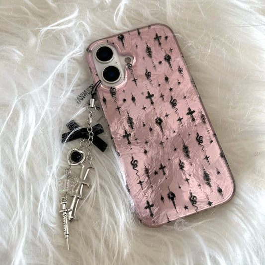 A pink phone case with a black gothic cross and skull pattern, featuring a dangling charm with a black ribbon bow, silver crossbones, and a small skull accent, displayed on a white faux fur background.