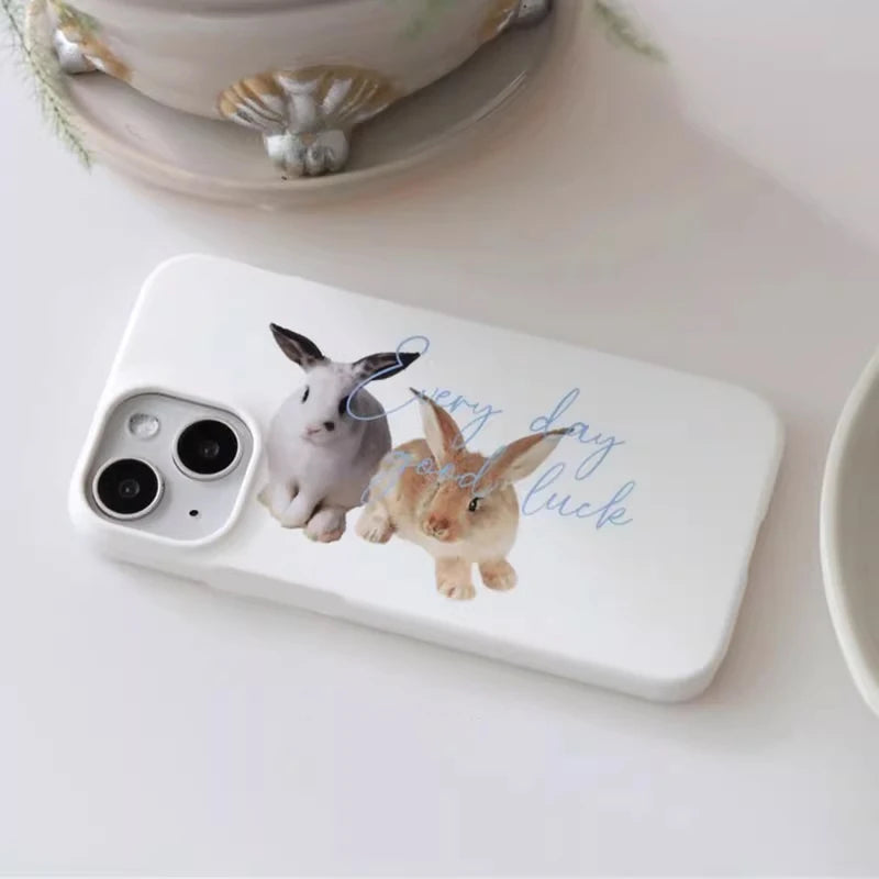 A white phone case featuring a printed design of two rabbits and the phrase 'Every day good luck' in light blue script, displayed on a light surface with a decorative bowl in the background.