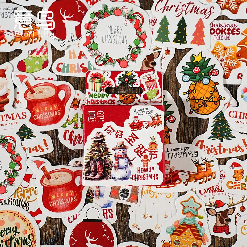 A collection of colorful Christmas-themed stickers scattered on a dark wood background, with a red and white gift box in the center. The stickers feature festive designs including Santa Claus, reindeer, Christmas trees, hot cocoa mugs, gingerbread houses, and holiday text such as 'Merry Christmas' and 'Howdy Christmas'.