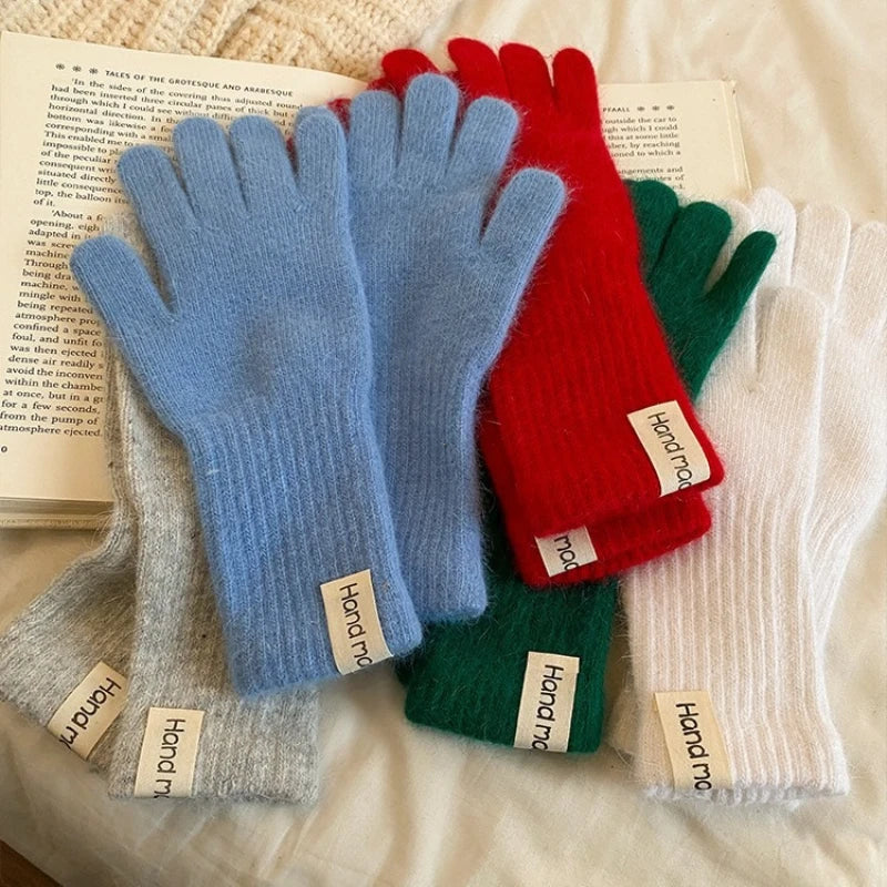 Four pairs of ribbed knit fingerless gloves in light blue, red, green, and white, arranged on an open book over a cream knit blanket. Each glove has a small white fabric tag reading 'Hand made'.