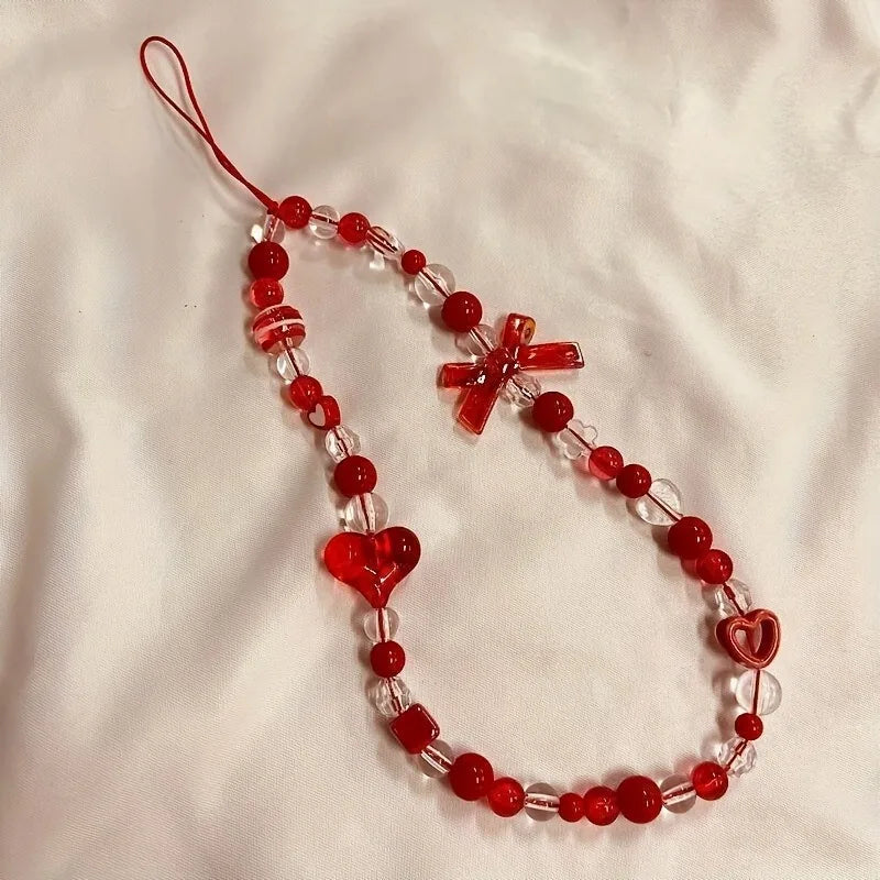 A red and clear beaded phone strap or bag charm laid flat on a white fabric background, featuring red beads, clear faceted beads, a red bow, a red heart, and a red heart charm.