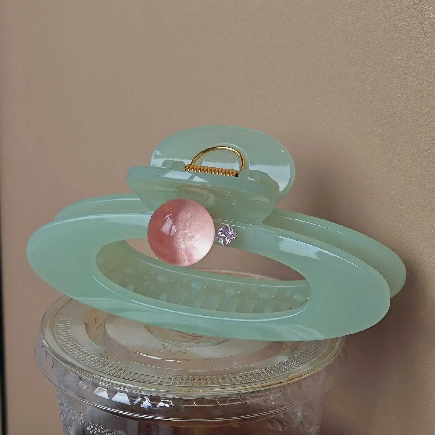 Light green hair claw with pink sphere and gold ring on a clear container against a beige background