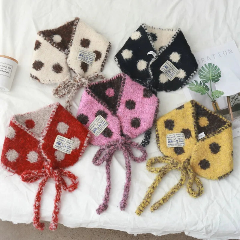 Five fuzzy knit bow neck warmers with polka dots are arranged on a white fabric background. The neck warmers come in red, pink, black, cream, and yellow, each with a matching tie bow.