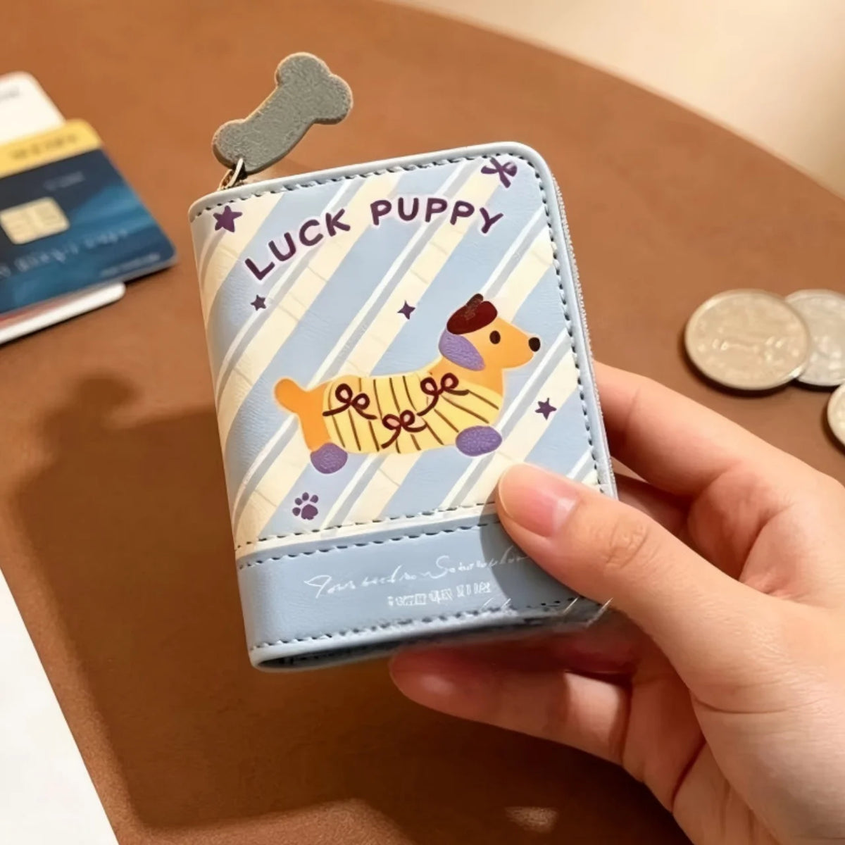 A hand holding a small zip-around wallet with a cartoon dog design and the words 'LUCK PUPPY' on a brown tabletop, with coins and cards blurred in the background.