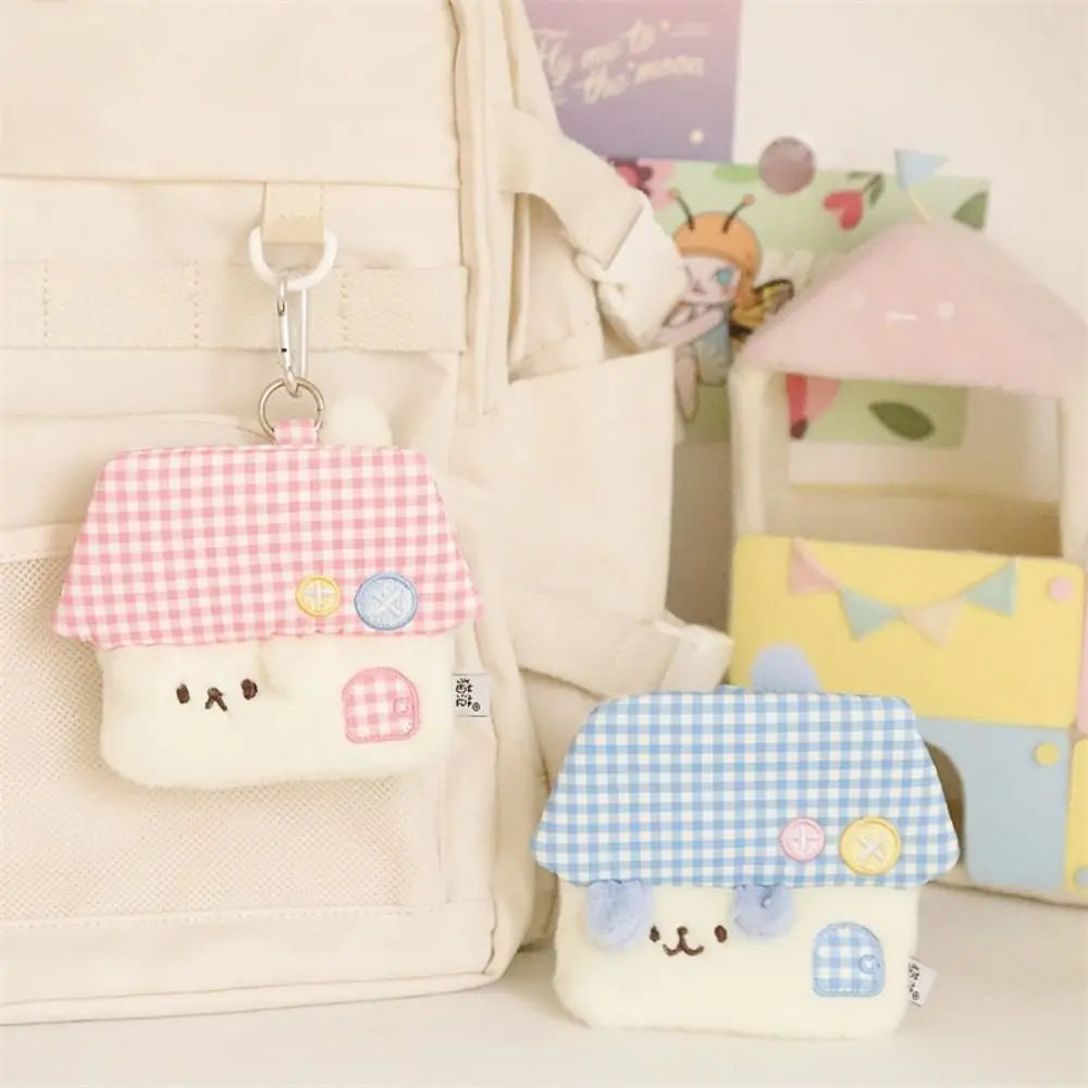 Two cute house-shaped coin purse keychains with embroidered faces, shown hanging on and next to a cream canvas backpack. One has a pink gingham roof, and the other has a blue gingham roof.