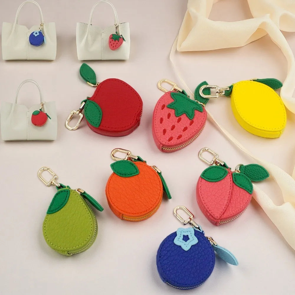 A collection of small, round, fruit-shaped zipper coin purses and keychain bags in red apple, pink strawberry, yellow lemon, green pear, orange, pink peach, blue blueberry, and white handbag designs, arranged on a light pink background with a cream ribbon.