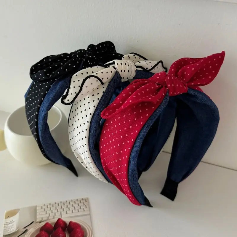 Three fabric headbands with large bow details are stacked on a white surface. The headbands feature black and white polka dots, white and navy blue polka dots, and solid navy blue with red polka dots.