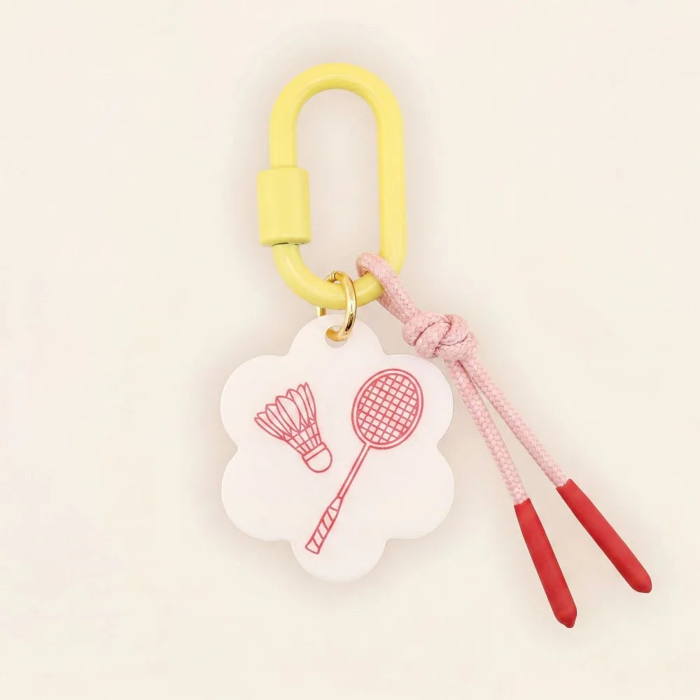 A yellow carabiner keychain with a white cloud-shaped charm printed with red badminton icons, attached to a pink braided cord with red tips, on a light cream background.